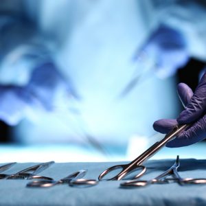 Nurse hand taking surgical instrument for group of surgeons at background operating patient in surgical theatre. Steel medical instruments ready to be used. Surgery and emergency concept